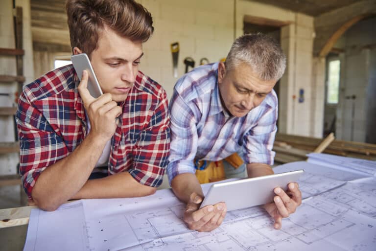 Un jeune homme en chemise à carreaux téléphone tandis qu'un homme plus âgé consulte une tablette numérique au-dessus de plans de construction étendus sur une table dans un chantier.