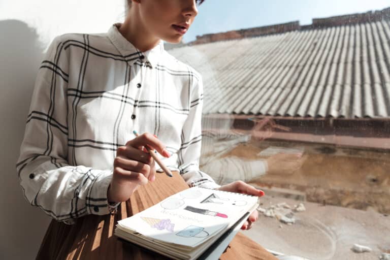 Une femme vêtue d'une chemise à carreaux dessine des accessoires de mode, tels qu'un rouge à lèvres et un sac à main, dans un carnet à croquis près d'une fenêtre.