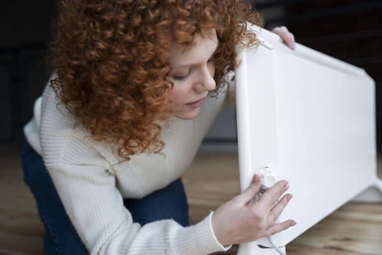 Une femme aux cheveux roux bouclés, vêtue d'un pull blanc, est agenouillée au sol et ajuste le thermostat d'un radiateur électrique portatif.