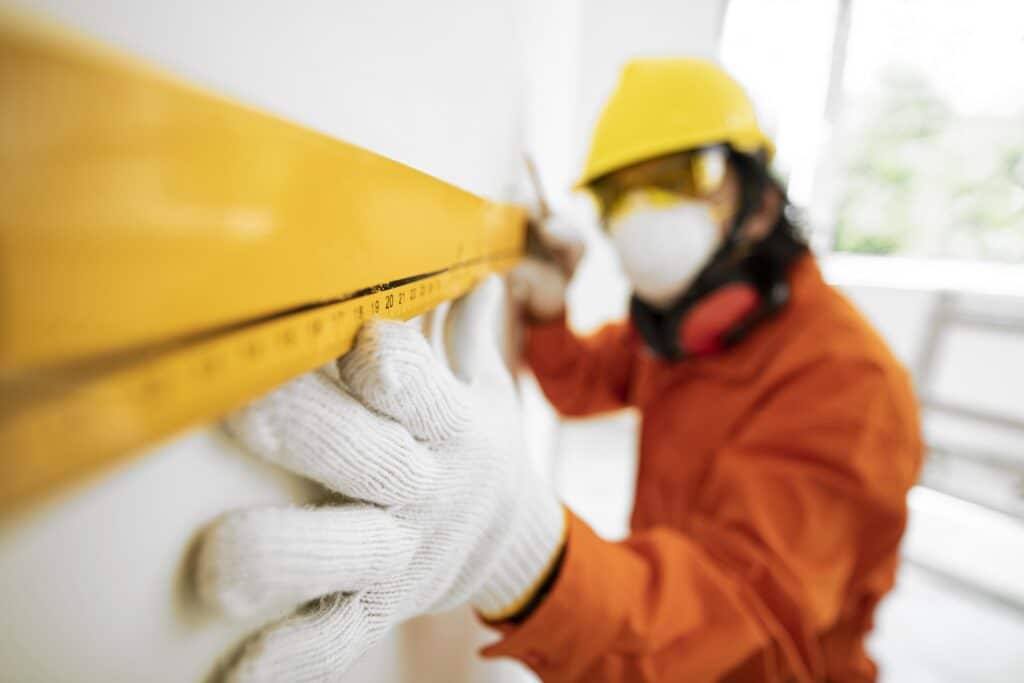 Un ouvrier portant un équipement de sécurité complet, incluant un casque jaune, un masque de protection et des gants, utilise une grande règle de maçon pour vérifier la planéité d'un mur.