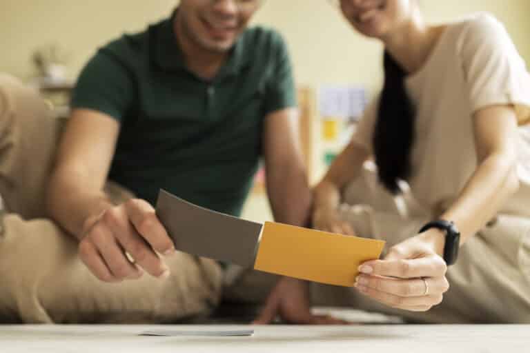 Un couple sourit en comparant des échantillons de peinture de couleur grise et jaune pour leur projet de décoration.
