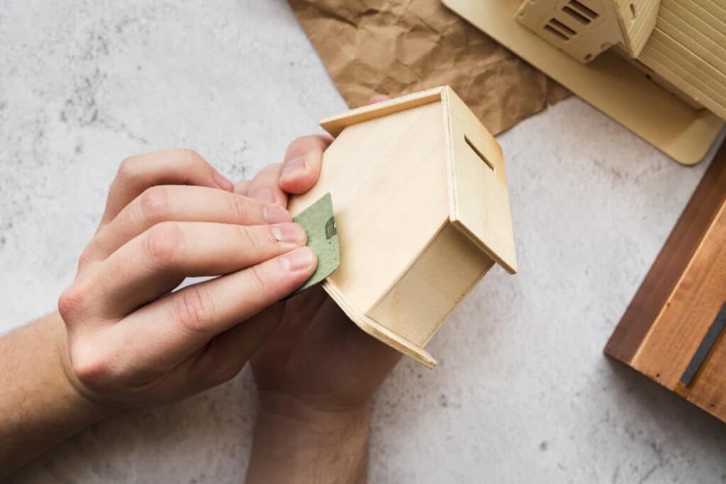 Une personne ponce les parois d'une petite maison miniature en bois à l'aide d'un morceau de papier de verre.