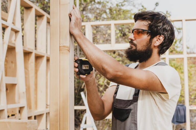 Un ouvrier barbu portant des lunettes de protection et une salopette prend les mesures d'une structure en bois avec un mètre ruban.
