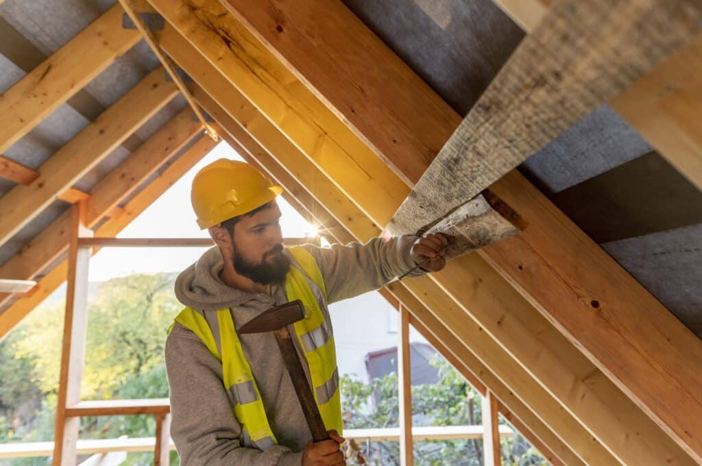 Un ouvrier en casque et gilet de sécurité travaille sur la charpente en bois d'un toit en construction.