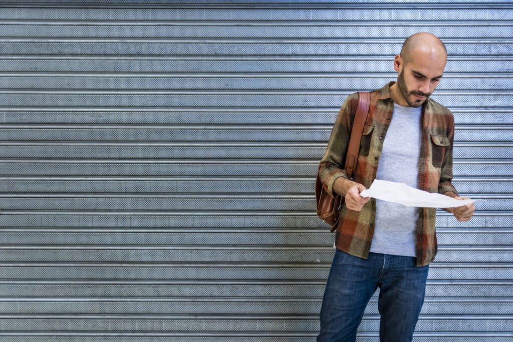 Un homme chauve avec une barbe, portant une chemise à carreaux, un t-shirt gris et un sac à dos, regarde un document devant une porte métallique à rideau roulant.