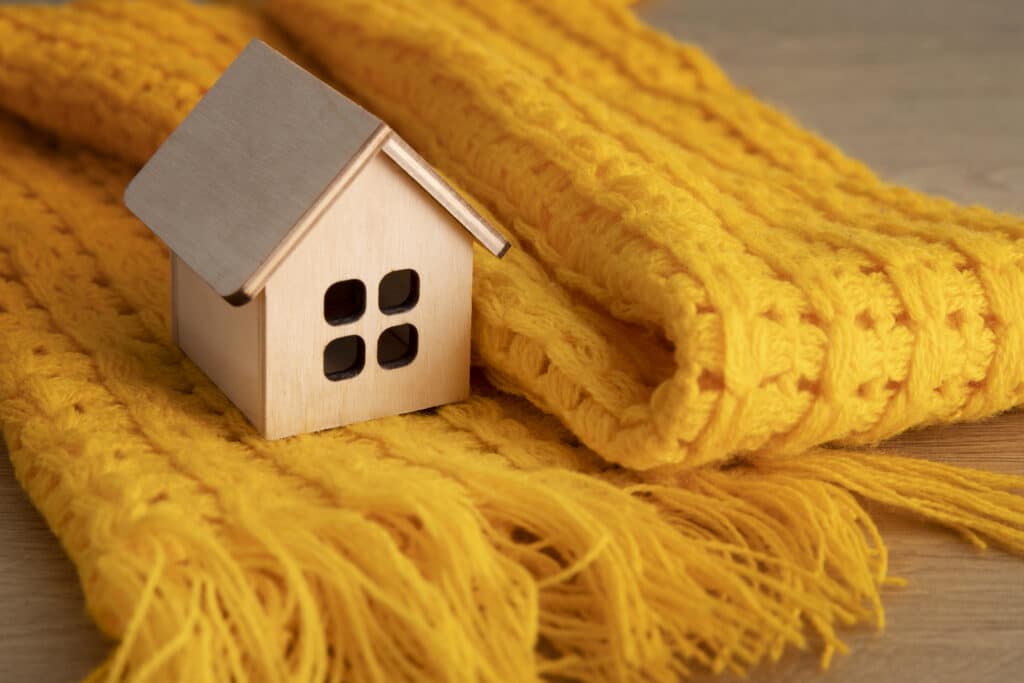 Une petite maison miniature en bois posée sur une écharpe en laine jaune épaisse, symbolisant le confort et la chaleur domestique.