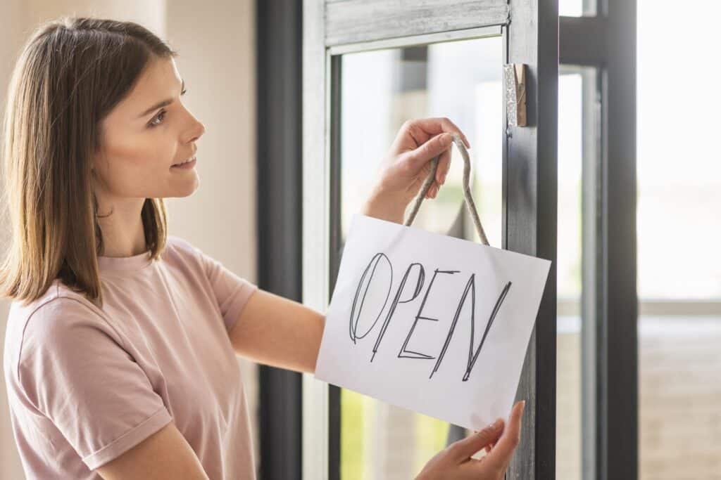 Une jeune femme souriante accroche un panneau blanc où est écrit "OPEN" à la main sur une porte vitrée.