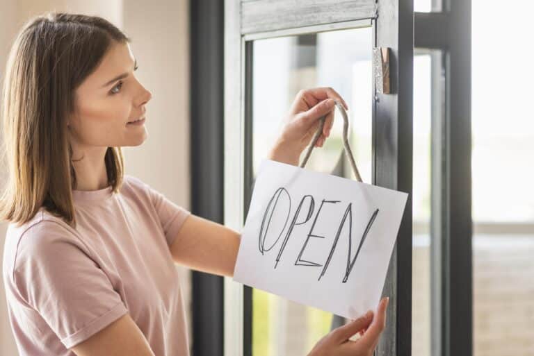 Une jeune femme souriante accroche un panneau blanc où est écrit "OPEN" à la main sur une porte vitrée.