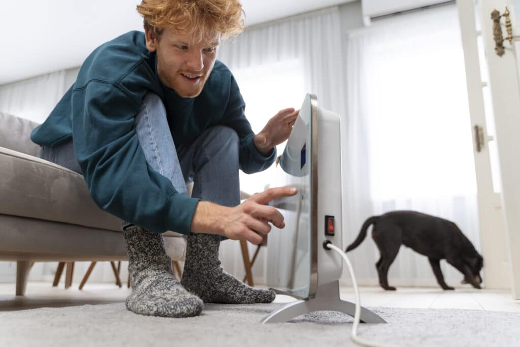 Un homme aux cheveux roux, assis sur un canapé et portant des chaussettes épaisses en laine, se penche pour régler ou allumer un radiateur électrique portatif moderne sur le sol. En arrière-plan, un chien noir marche près d'une fenêtre lumineuse.