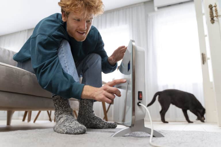 Un homme aux cheveux roux, assis sur un canapé et portant des chaussettes épaisses en laine, se penche pour régler ou allumer un radiateur électrique portatif moderne sur le sol. En arrière-plan, un chien noir marche près d'une fenêtre lumineuse.