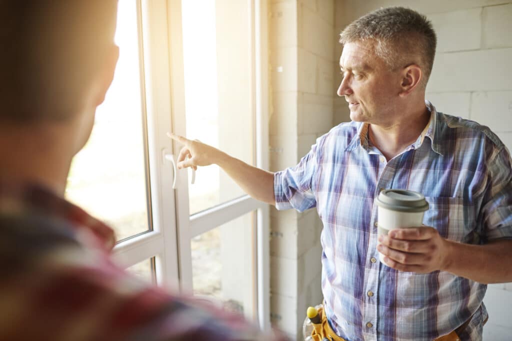 Un homme portant une chemise à carreaux et une ceinture porte-outils tient un gobelet de café tout en montrant une fenêtre à une autre personne dans une pièce en cours de construction.