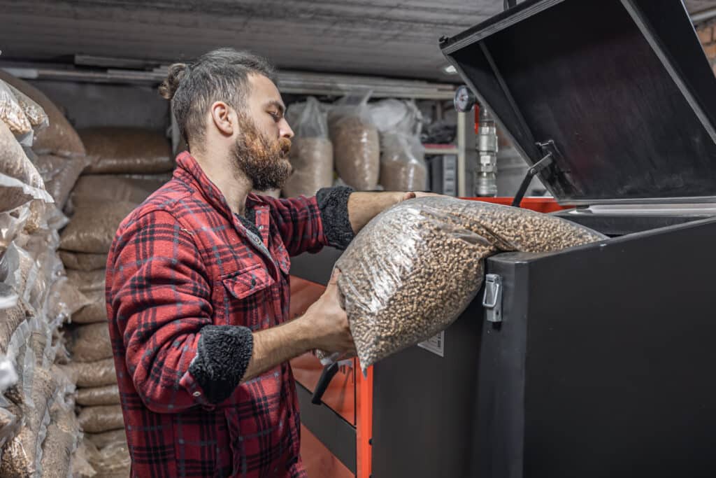 Un homme barbu portant une chemise à carreaux rouges remplit une chaudière avec un sac de granulés de bois, dans un local de stockage rempli de nombreux autres sacs similaires.