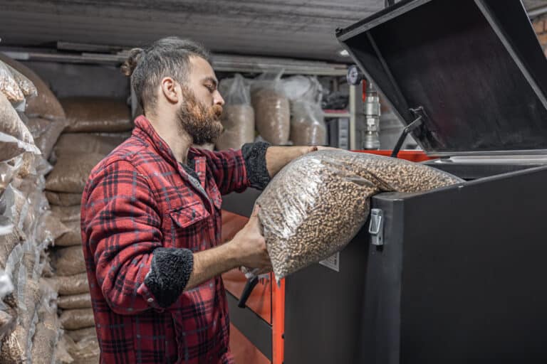 Un homme barbu portant une chemise à carreaux rouges remplit une chaudière avec un sac de granulés de bois, dans un local de stockage rempli de nombreux autres sacs similaires.