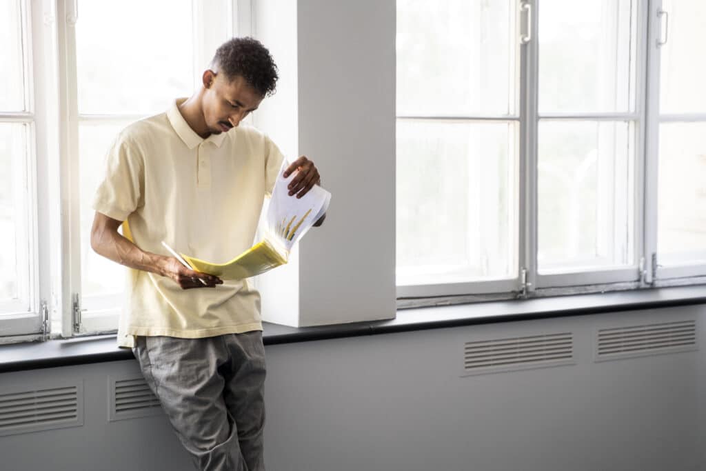 Un homme en polo jaune, appuyé contre le rebord d'une fenêtre dans un espace lumineux, examine attentivement des documents de travail.