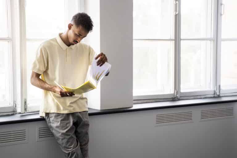 Un homme en polo jaune, appuyé contre le rebord d'une fenêtre dans un espace lumineux, examine attentivement des documents de travail.