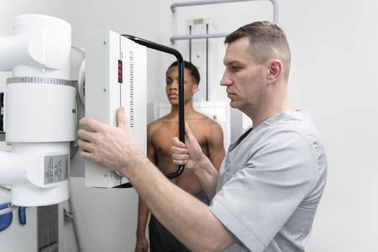 Un manipulateur en radiologie ajuste l'équipement d'imagerie médicale devant un jeune homme torse nu qui se tient prêt pour une radiographie.