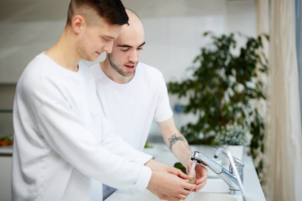 Deux jeunes hommes en t-shirts blancs sont debout dans une cuisine lumineuse et lavent un fruit sous le robinet de l'évier.