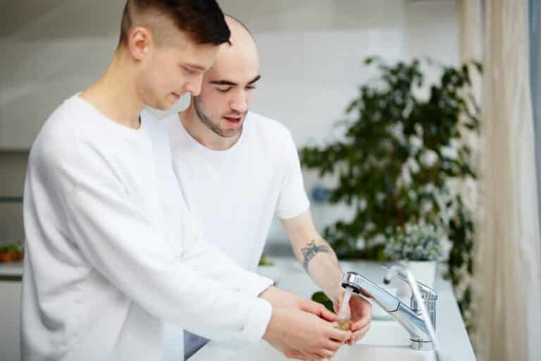 Deux jeunes hommes en t-shirts blancs sont debout dans une cuisine lumineuse et lavent un fruit sous le robinet de l'évier.