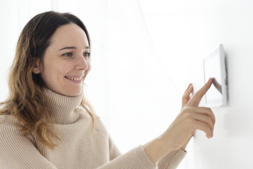 Une femme souriante portant un pull à col roulé beige utilise son doigt pour régler un thermostat numérique fixé sur un mur blanc.