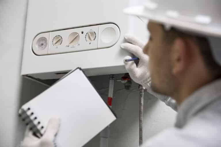 Un technicien portant un casque de chantier et des gants blancs inspecte une chaudière murale tout en prenant des notes sur un carnet.