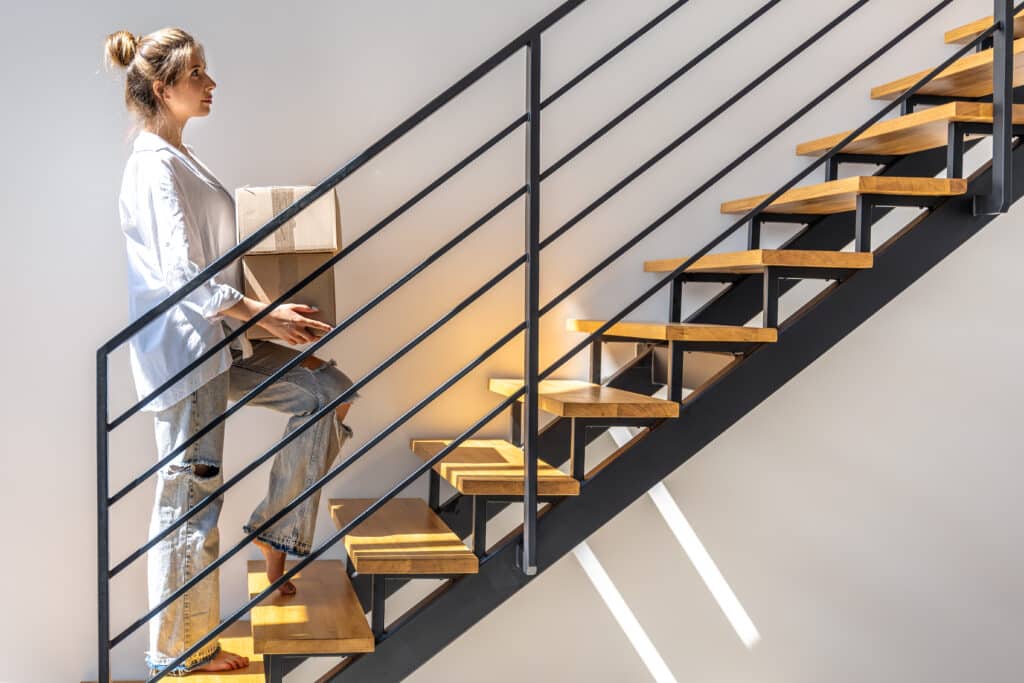 Une jeune femme portant une chemise blanche et un jean monte pieds nus un escalier moderne en bois et métal tout en tenant un carton dans ses bras.