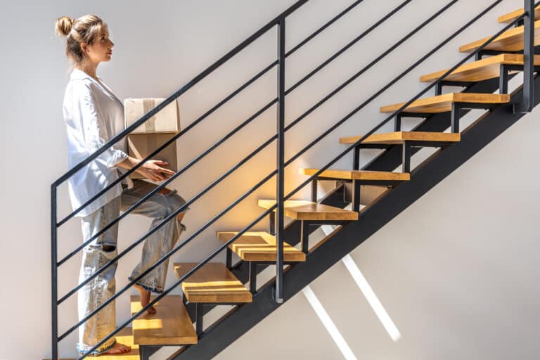 Une jeune femme portant une chemise blanche et un jean monte pieds nus un escalier moderne en bois et métal tout en tenant un carton dans ses bras.
