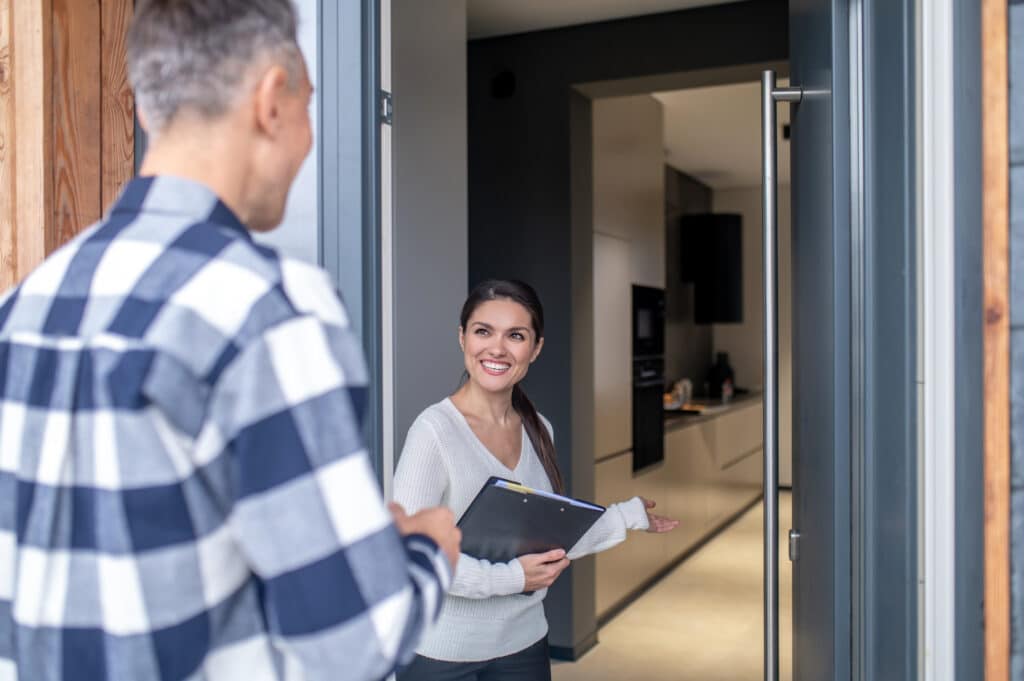 Une femme souriante tenant un dossier accueille un homme à l'entrée d'une maison en lui faisant un geste de la main pour l'inviter à entrer.